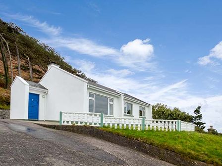 A white house with a blue door on a hill with trees and grass at Rossbeigh Beach Cottage No 6 Rossbeigh near Glenbeigh County Kerry