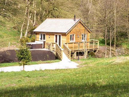 A wooden cabin with a deck and hot tub in a grassy and wooded area at Park Brook Retreat in Scorton