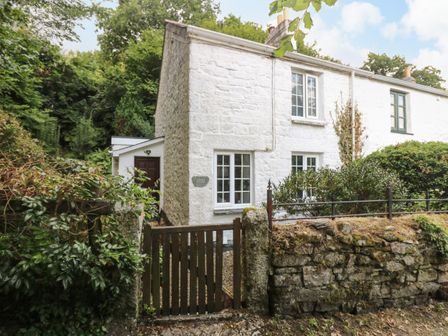 A white stone cottage with windows a wooden gate and stone wall surrounded by greenery at Tinners Cottage in Liskeard