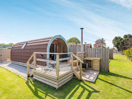 An outdoor pod accommodation with a small deck balcony and two chairs surrounded by grass at Two Hoots in Northop