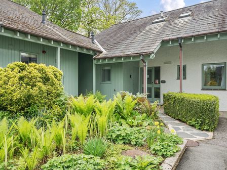 The exterior of a cottage with green wooden panels white walls a slate roof and a garden with various plants and bushes at Elm - Woodland Cottages in Bowness-on-Windermere