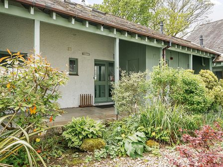 An exterior view of a building entrance with green door and wooden paneling surrounded by various bushes and plants at Cherry - Woodland Cottages in Bowness-in-Windermere