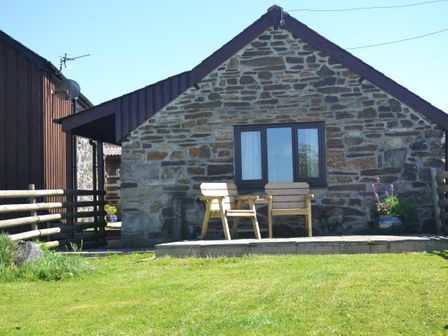 A stone cottage with two wooden chairs and a small table on a patio with a grassy lawn at Nestling Barn Peter Tavy near Tavistock