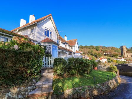 A white house with a tiled roof and a garden with green bushes at 2 Moorlands in Minehead