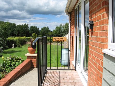A side entrance to a brick house with a black metal gate with a Beechnt Cottage sign and a garden with green grass and trees in the background at Beechnut Cottage in Tewkesbury