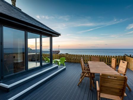 An outdoor deck with wooden table and chairs and green chairs overlooking the sea at Rocky Close in St Ives