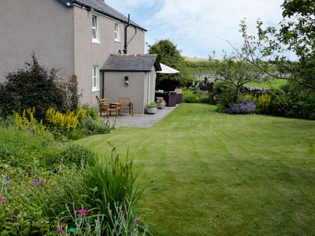 A garden with lawn, flower beds, trees, wooden chairs, and a table with umbrella outside a house at Waingate Cottage in Flookburgh