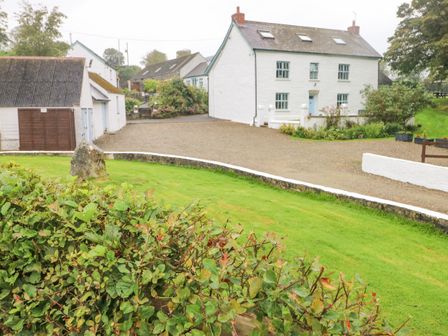 A white house with blue windows and doors next to a garage with brown doors and a gravel courtyard surrounded by green grass and bushes at Y Cartws in Nevern near Cardigan