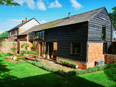 An exterior view of a two-story black wooden and brick building with a garden patio and lawn at The Hayloft in Ross-on-Wye