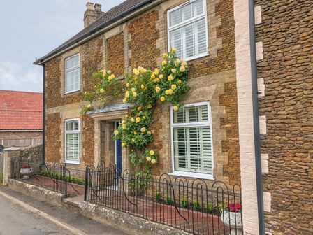 A stone house with white shutter windows and yellow flowers climbing around the door at Bertelle in Grimston