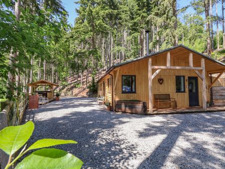 A wooden cabin with a small porch and bench surrounded by tall trees and gravel ground at Lady Bagots Cabin in Ruthin