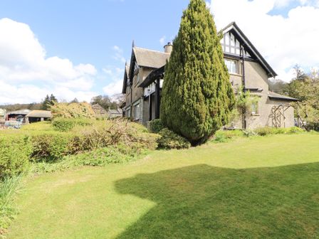 A grassy garden area with bushes and a tall tree next to a house with a slate roof at Bishop Howe in Far Sawrey