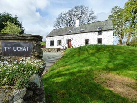 A white cottage with a slate roof and chimney behind a grassy lawn with trees and a stone sign that says Ty Uchaf in Llangwm near Bala