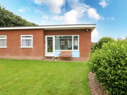A brick house with a white door and windows a wooden bench a green lawn and bushes at 20 Siesta Mar in Mundesley