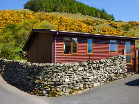 A wooden cabin with windows behind a stone wall on a paved road with hills and trees in the background at South Lodge in Kendal