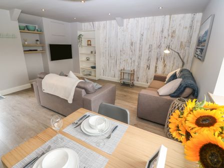 A living room with two sofas a wall-mounted TV a dining table set with glasses and plates and sunflowers on the table at Bodsela Bach in Nefyn