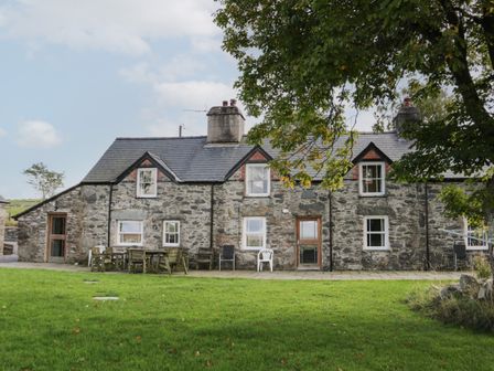 A stone farmhouse with multiple windows and doors a patio area with outdoor tables and chairs and a large tree with green leaves in front at Bryn Dedwydd Farmhouse in Betws-y-Coed