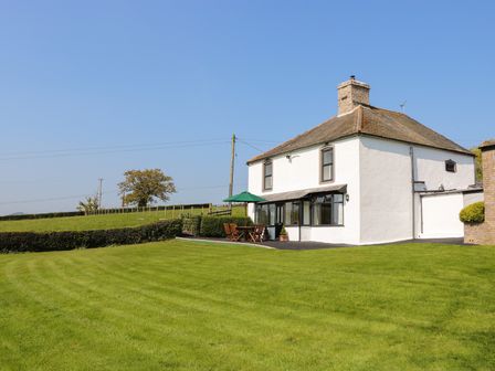 A white two-story house with a chimney sitting on a large green lawn with outdoor furniture and an umbrella at Cefn Isaf in Betws yn Rhos near Abergele