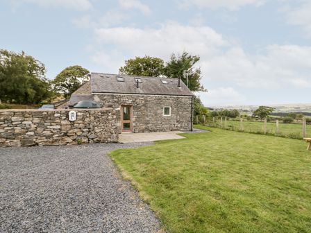 A stone cottage with a slate roof next to a gravel driveway and a grass lawn with a wooden fence in the background at Bryn Dedwydd Cottage in Pentrefoelas