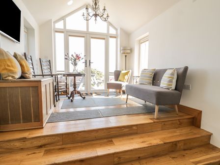 A sitting area with wooden flooring steps a grey sofa and armchair a round wooden table with flowers and glass doors at Marsh Villa in Silloth