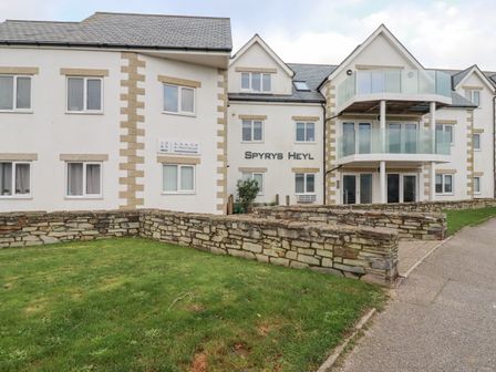A white multi-story building with stone accents and balconies surrounded by grass and stone walls at Spyrys Heyl in Newquay