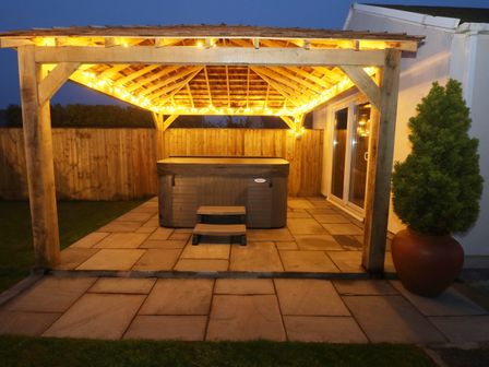 A wooden gazebo with lights surrounding a hot tub on a stone patio with a potted plant on the side at The Elms in Milford Haven