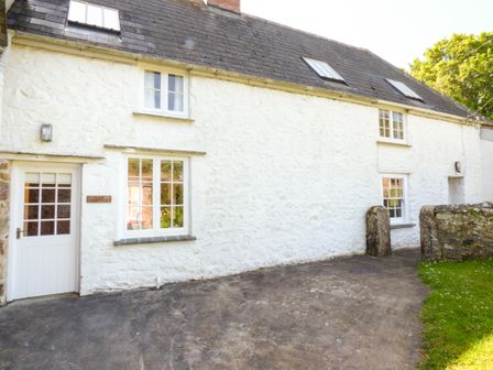 A white stone cottage with windows and a door next to a driveway and grassy area at Farm Cottage in Marazion