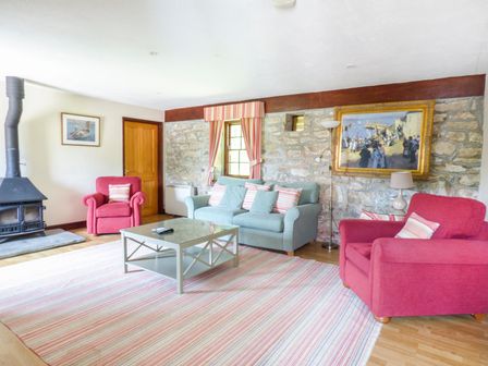 A living room with two red armchairs a blue sofa a glass coffee table a striped rug a wood burning stove and a stone wall at Flower House in Marazion
