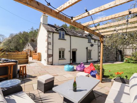 An outdoor patio area with a pergola, seating including sofas and ottomans, colorful bean bags, a table with a bottle and glasses, and a white stone house in the background at Nant Y Celyn in Clocaenog near Clawdd-Newydd