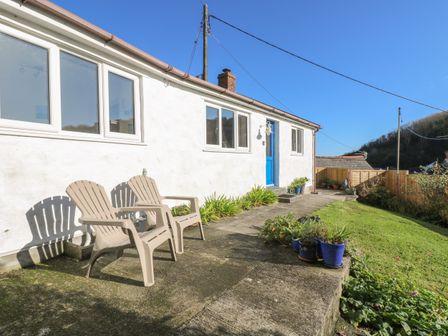 The exterior of a white house with a blue door two plastic chairs on a patio and several potted plants at Cockle Island Cottage in Porthallow