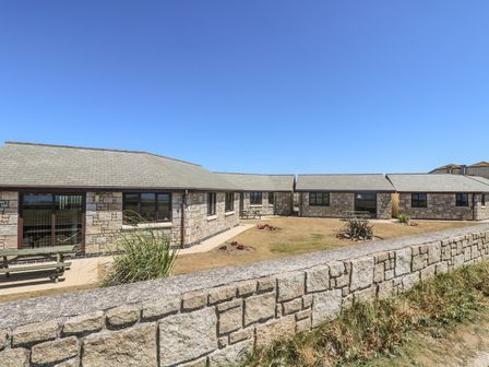Stone buildings with multiple windows and picnic tables in a grassy courtyard behind a low stone wall at Sea Spray in Marazion