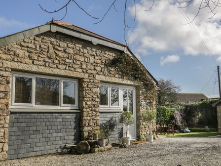 A stone building with white framed windows and door with potted plants outside and a garden seating area at Honeysuckle Cottage in Helston