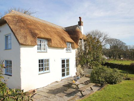 A cottage with a thatched roof and blue windows with a picnic table bench and garden bench outside Rose Cottage in Manaccan