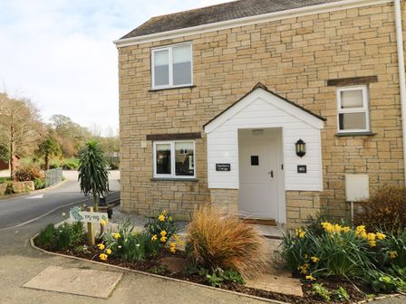 The entrance of a stone house with a white door and porch surrounded by flower beds with yellow flowers at Heather's Cottage in Falmouth
