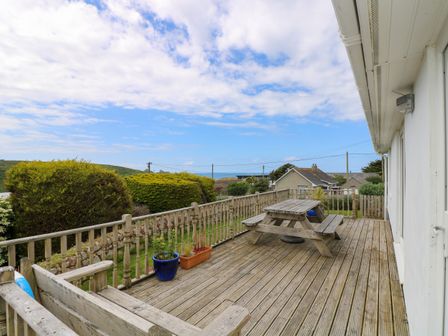 An outdoor wooden deck with a picnic table potted plants and railing overlooking bushes and houses at Bethany Bay View in Holywell Bay