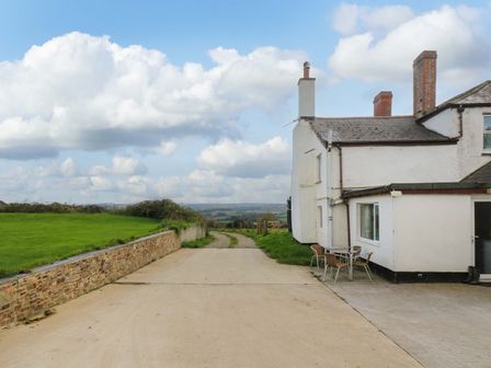 An exterior view of a white house with chimneys and outdoor table and chairs on a paved area near a concrete driveway with green grass and a stone wall at The Cottage in Bideford