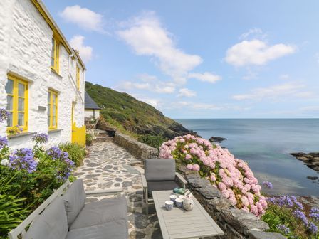 An outdoor patio with gray seating and a table overlooking the sea next to a white building with yellow windows and a stone pathway at Kerbenetty Harbour Cottage in Portloe