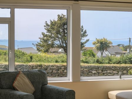 A seating area with a gray armchair and window showing trees houses and the sea at Seaport Lodge in Mawgan Porth