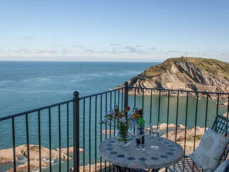 A balcony with a round table holding a vase of flowers and two glasses overlooking the sea and rocky coastline at Seascapes in Ilfracombe