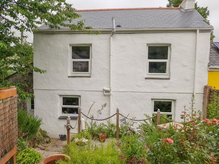 A white two-story house with four windows a small garden with plants and flowers and a metal chair at The Old Sweet Shop in St Blazey