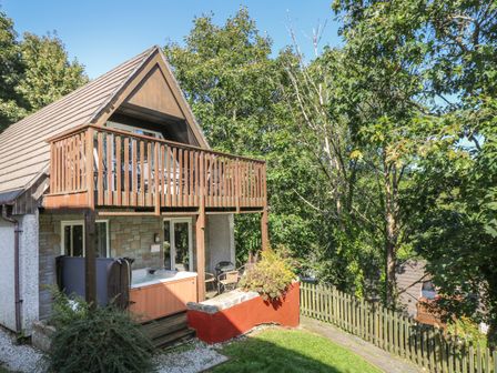 A two-story cabin with a wooden balcony and outdoor hot tub surrounded by trees at Valley Lodge 58 in Callington