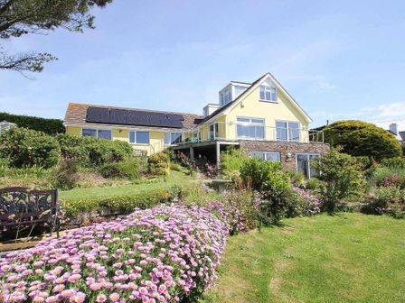 A yellow house with solar panels on the roof surrounded by a garden with pink flowers at Windrush in Mawgan Porth