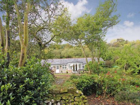 A stone cottage surrounded by trees and shrubs with a picnic bench in front at Dairy Cottage in Bodmin