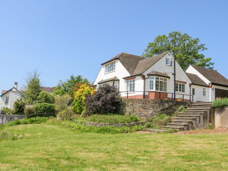 A white house with a sloped roof and a stone retaining wall in front of a grassy lawn at Charlesworth in Braunton
