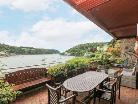 An outdoor patio with a wooden dining table and chairs overlooking a river with boats at Bight Boathouse in Dartmouth