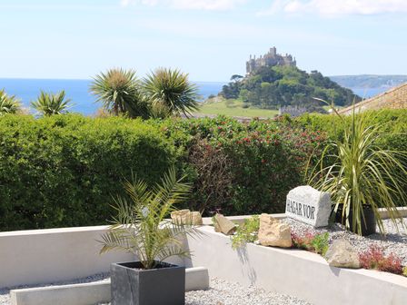 A garden with potted plants and a stone sign reading HAGAR VOR with a castle on a hill and the sea in the background at Hagar Vor in Marazion