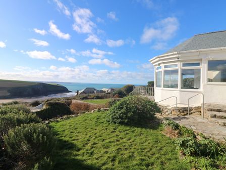 A house with large windows and a stone patio overlooking a garden with shrubs and a view of the sea at Gwillen in Mawgan Porth
