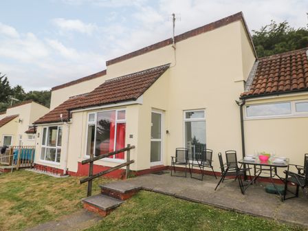 An exterior view of a house with a small patio, outdoor chairs and table set, and a grass lawn at Manorcombe 17 in Callington