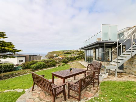 An outdoor patio area with wooden benches and table near a modern house overlooking a coastal landscape at Little Trelawns in Mawgan Porth