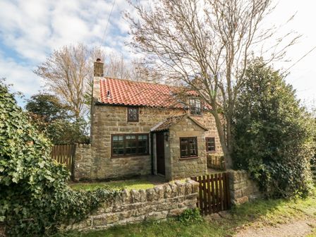 A stone cottage with a red tiled roof behind a low stone wall with a wooden gate and trees around at Holly Cottage in Whitby